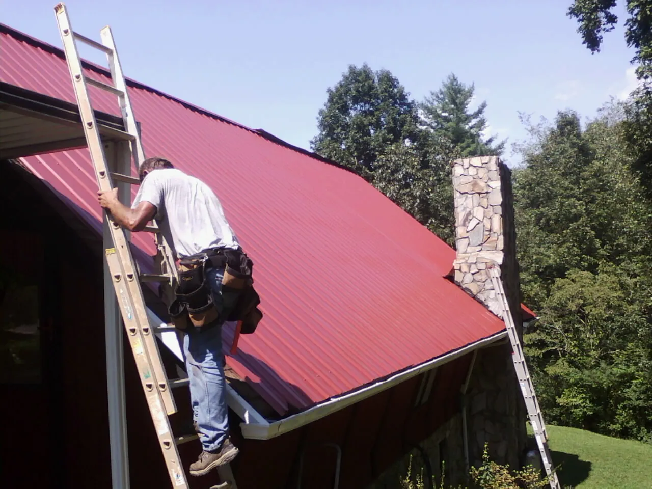 Roofer on a ladder beside a red metal roof and stone chimney — Soffit & Fascia Repair by Roofing Brothers in Newville